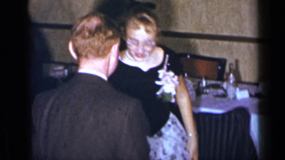 The woman smiles during the dance, wearing glasses, a black dress and pearl necklace as the suited man faces away from the camera.