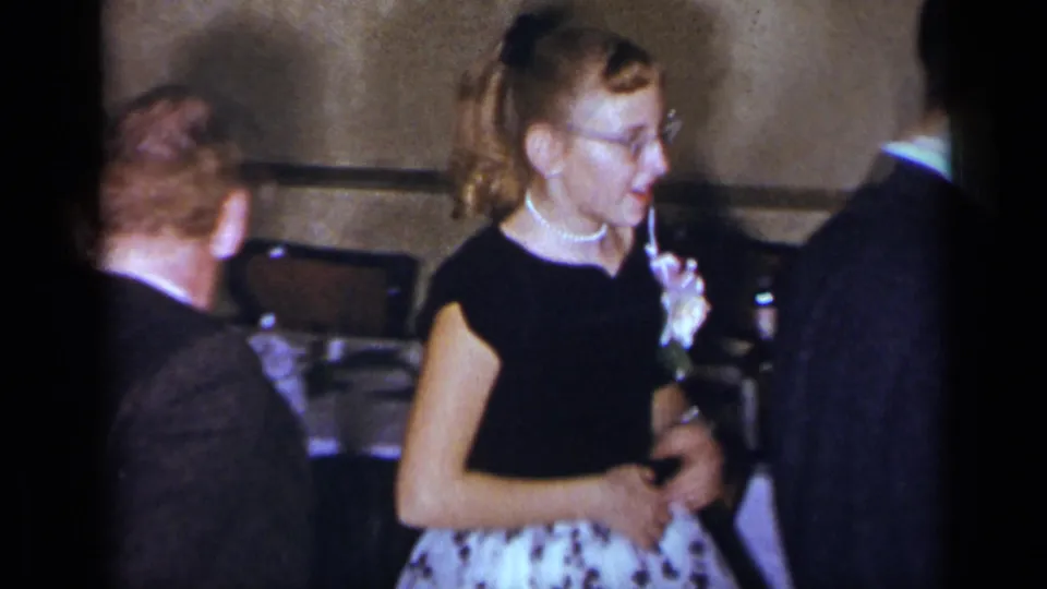 The woman in glasses and pearls appears between suited men in the 1954 New York interior, with dining tables behind her.