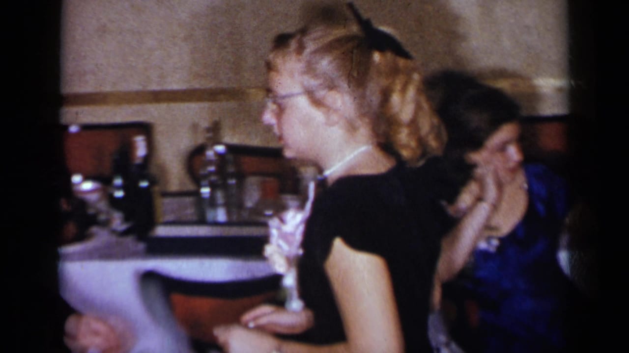 Young woman in glasses holds a corsage while standing near tables and seated guests.