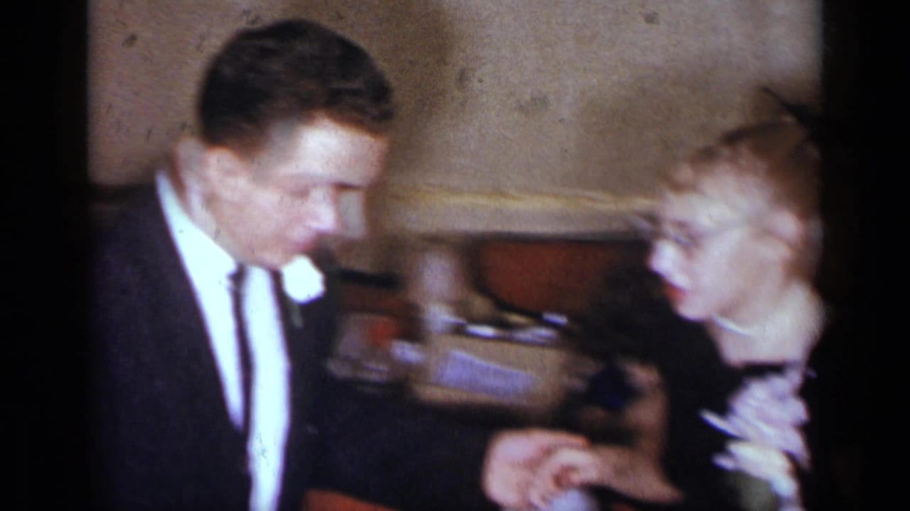 A suited man with a boutonniere faces a young woman in glasses as they dance closely in a dim indoor venue.