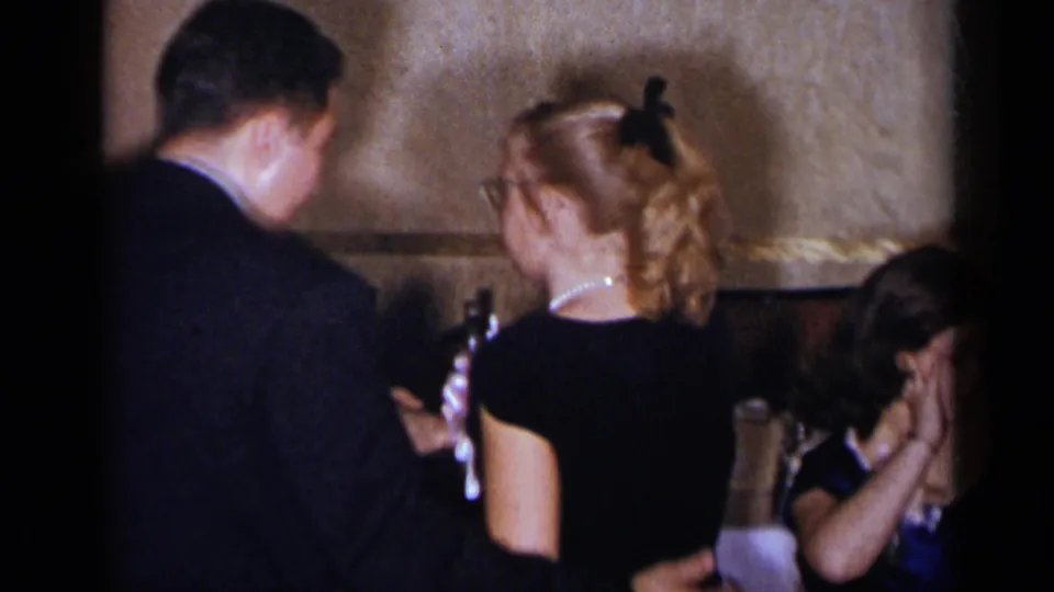 The couple is viewed from behind in the indoor venue, with the woman’s ponytail and nearby seated guest visible.