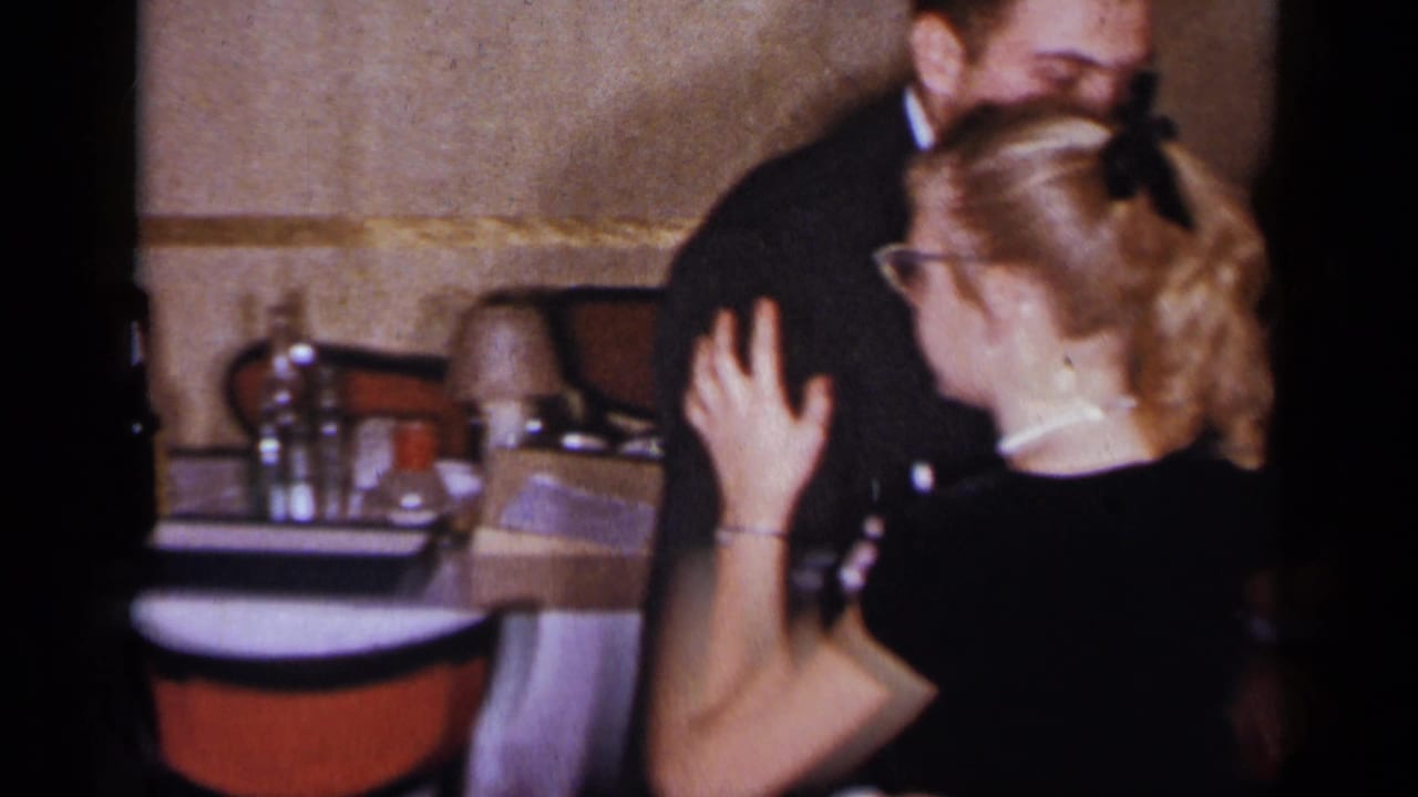 Couple dancing closely beside restaurant tables with red chairs.