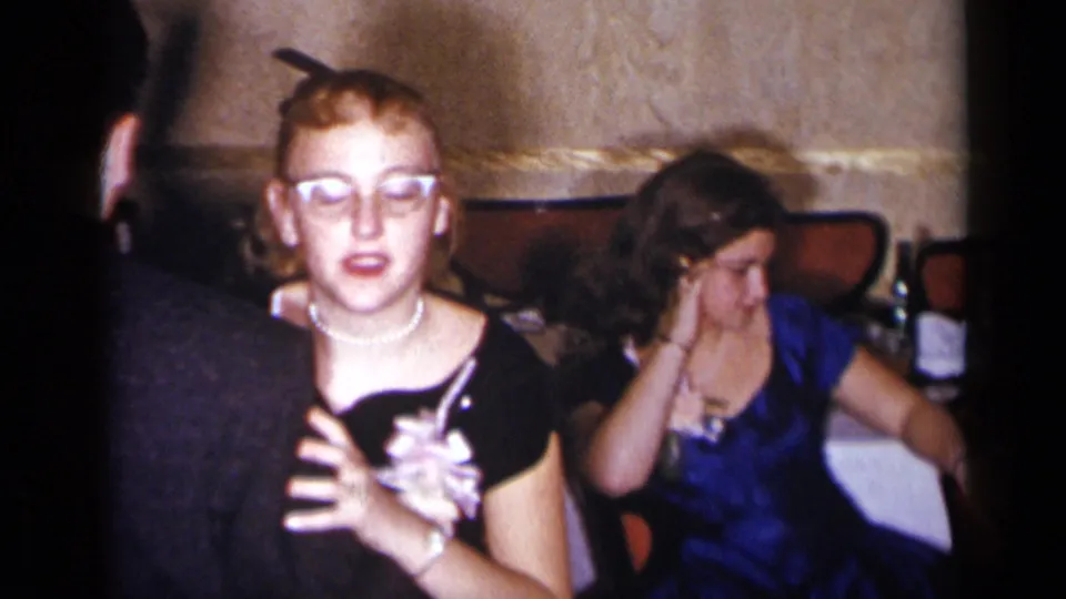 A woman with glasses holding a drink stands near a seated woman in a blue dress at a 1954 New York reception.