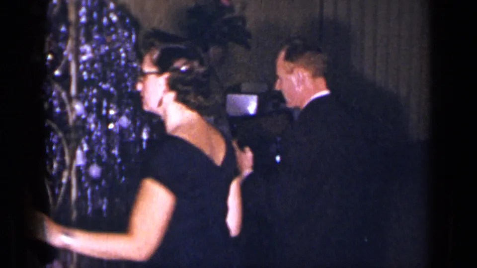 1954 New York home-movie frame of a woman in glasses and a black dress dancing near a tuxedoed man and shiny decorations.