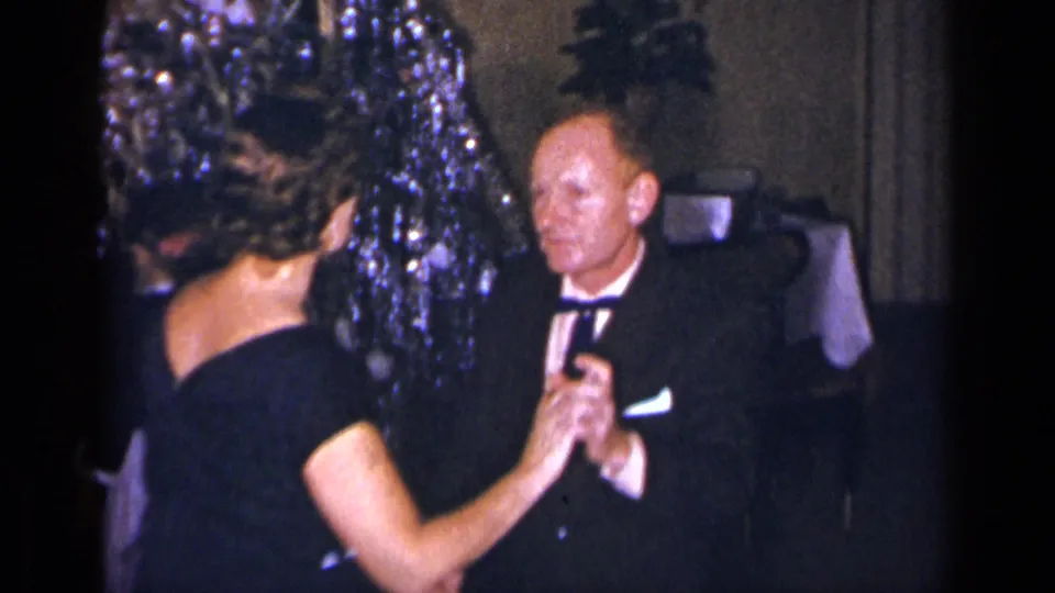 1954 New York frame showing a woman in a black dress dancing with a man in a tuxedo in a decorated interior.