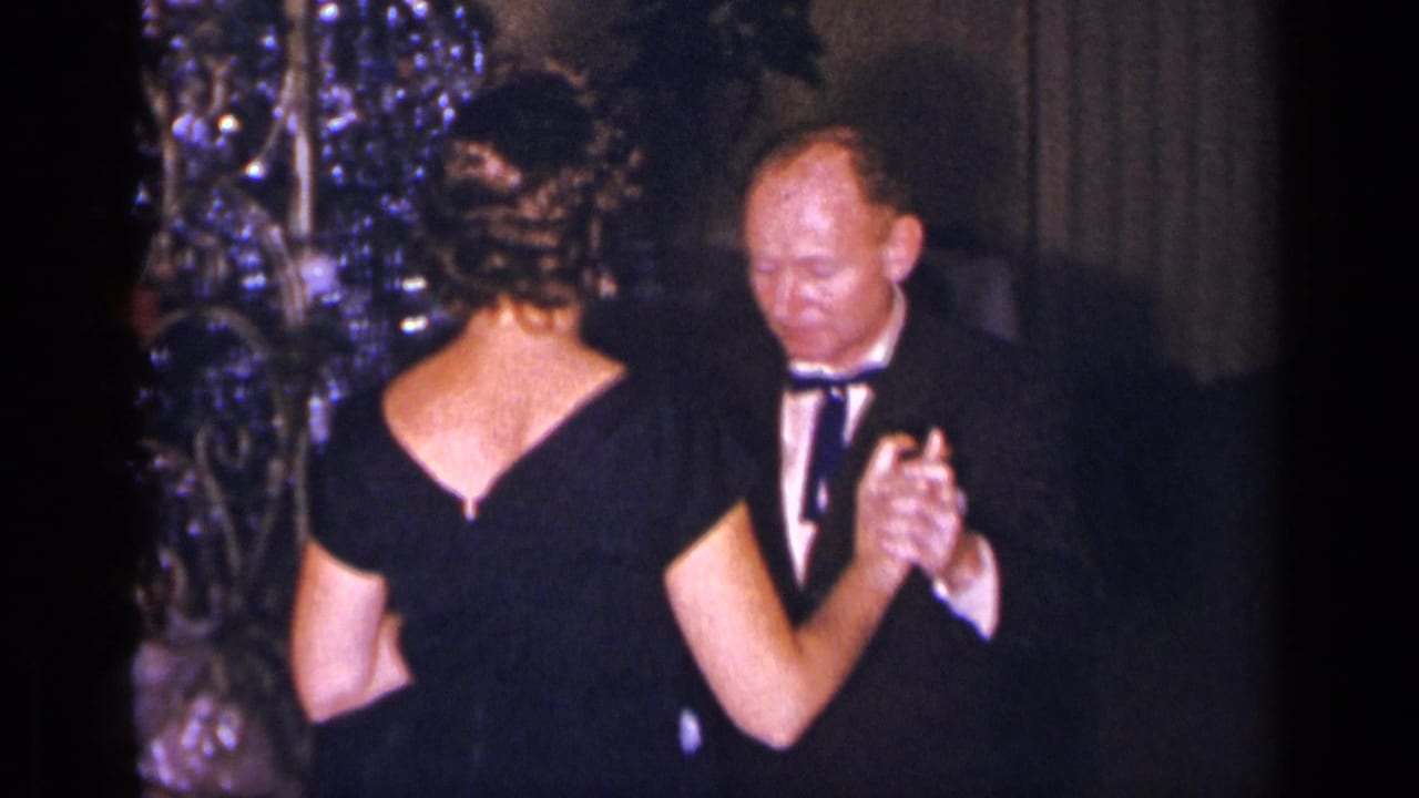 Man in a tuxedo dances with a woman in a black evening gown beside a decorated interior tree.