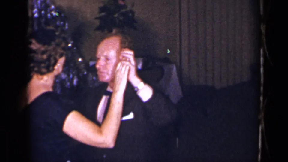 The formally dressed couple continues dancing in a private 1954 New York interior, framed closely by the home-movie camera.