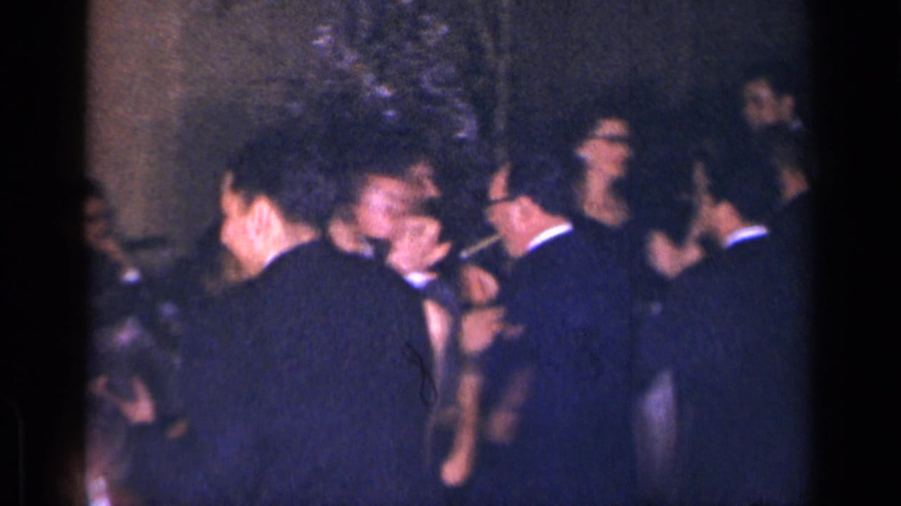 Adults in formal clothing gather and dance in a dim indoor room.