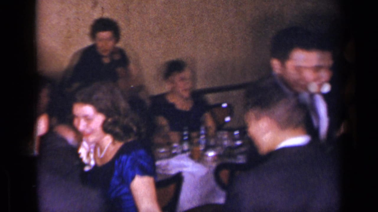 Seated and standing guests gather beside a banquet table with glasses.