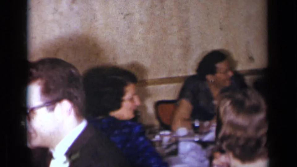 1954 New York home-movie frame showing guests seated near a banquet table, with others conversing along the wall.