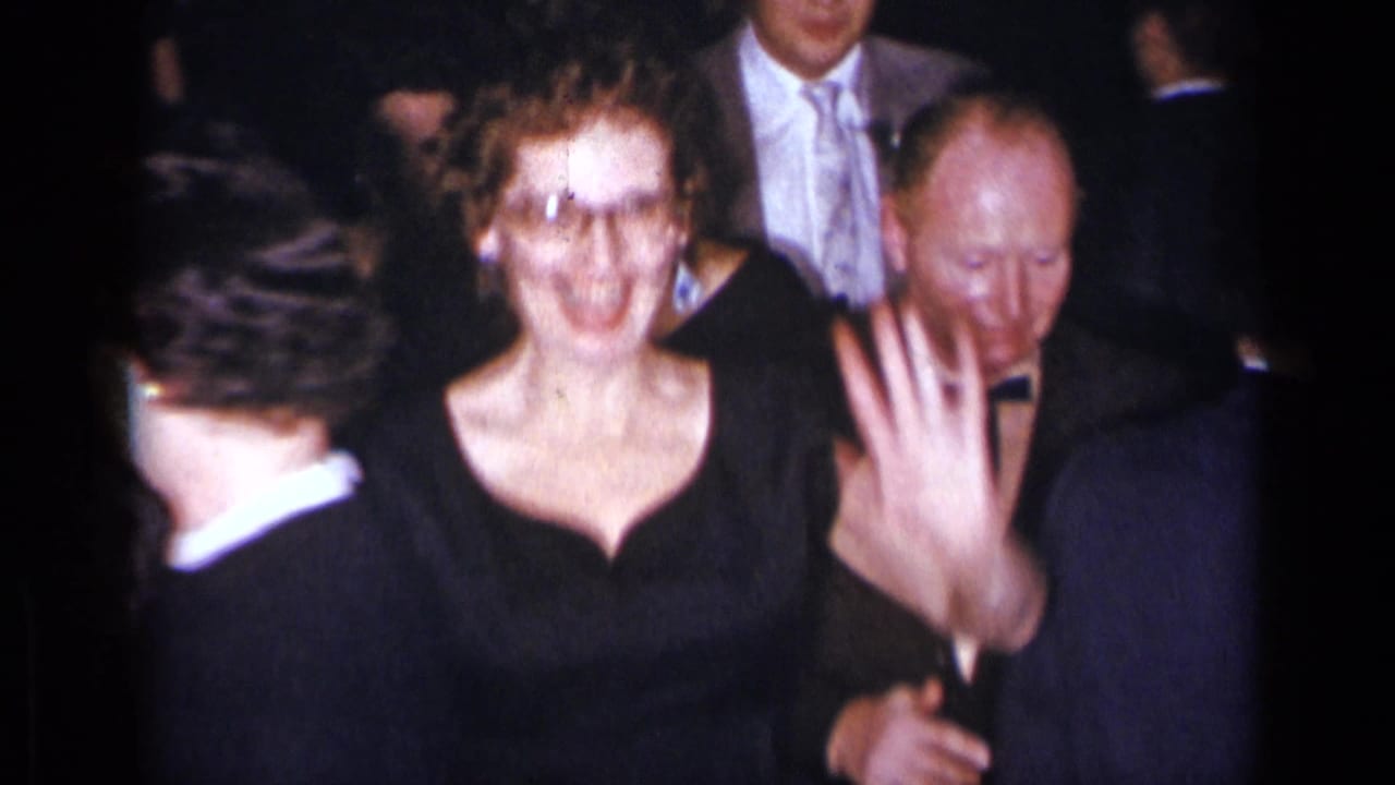 Smiling woman in black dress and glasses waves among men in suits.