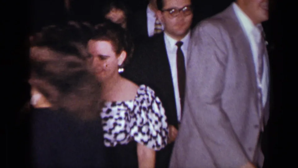 Formal reception guests in suits and patterned dress pass through a dim interior in this 1954 New York home-movie frame.