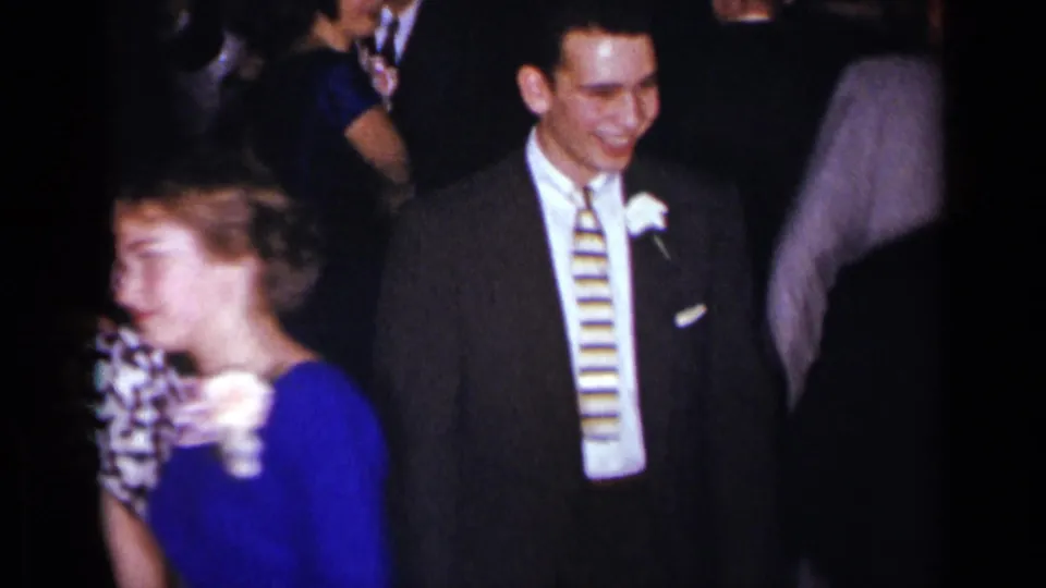 Smiling man with boutonniere and striped tie stands near a woman in blue at a 1954 New York formal gathering.
