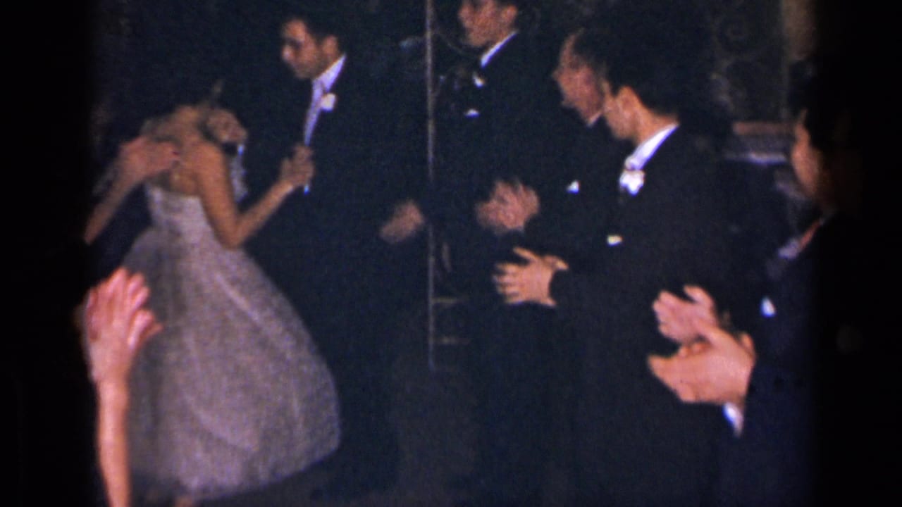 Woman in a white gown dances as men in tuxedos and guests clap nearby.