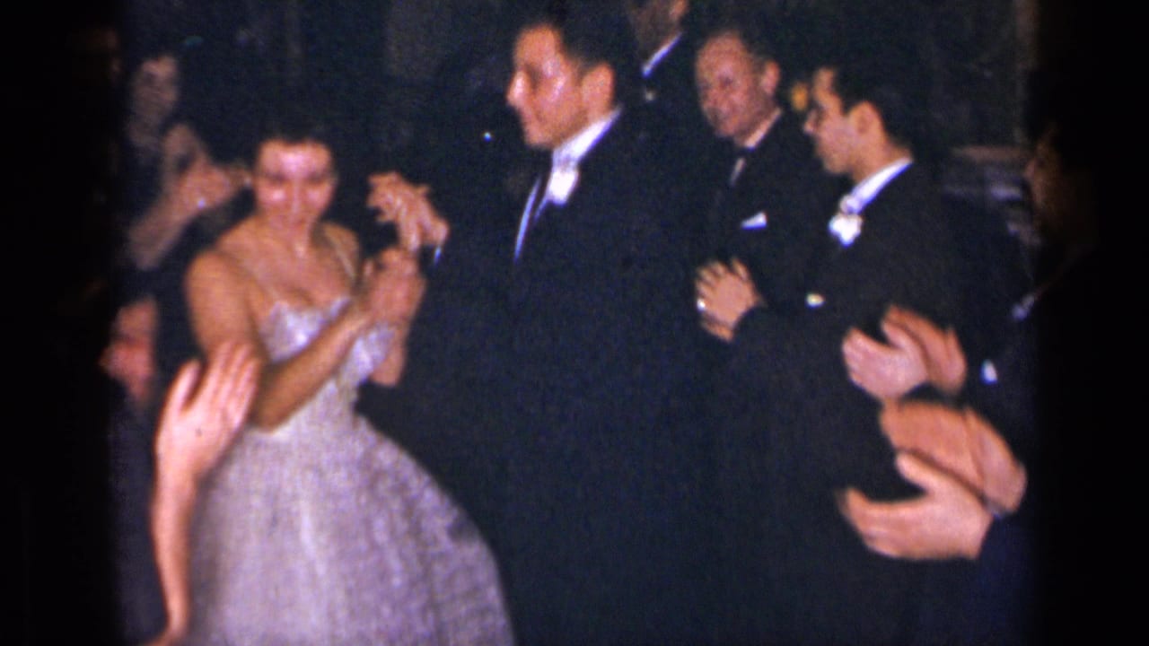 Woman in a white gown dances with a man in a tuxedo while guests clap.
