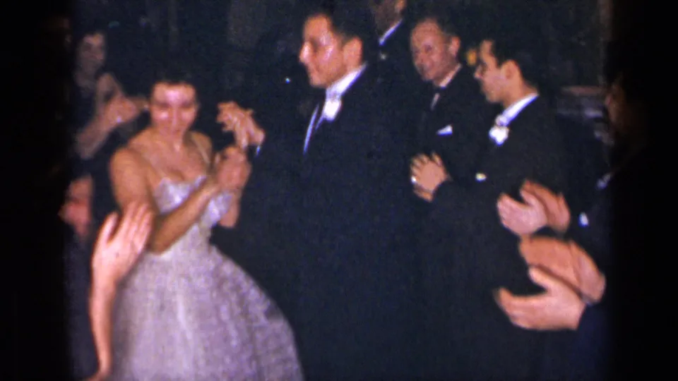 A woman in a white gown dances with a tuxedoed man as surrounding formal guests clap in dim 1950s home-movie footage.
