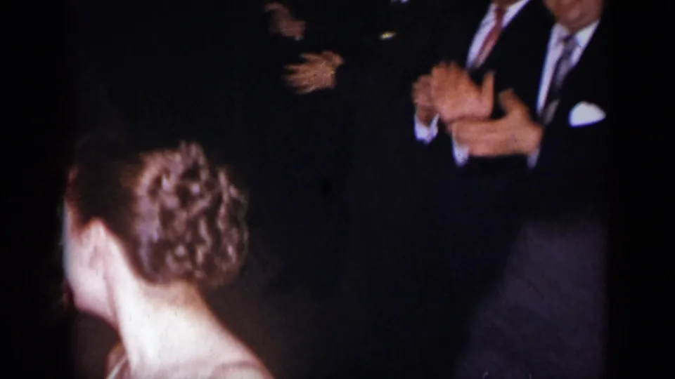 The camera catches the back of a woman's styled hair in the foreground while formally dressed guests clap in a dark reception setting.
