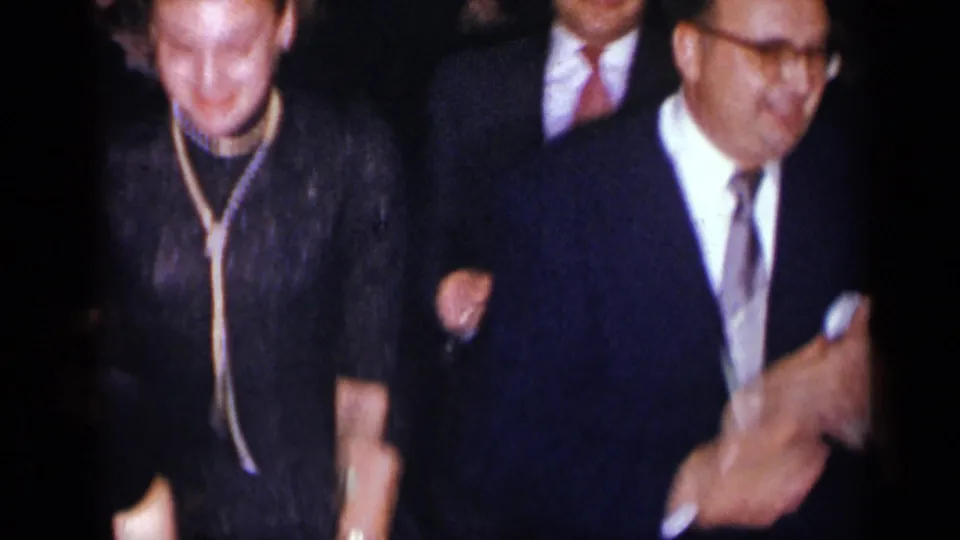 An older woman in a dark dress and a man in a suit pass close to the camera during a crowded formal celebration.
