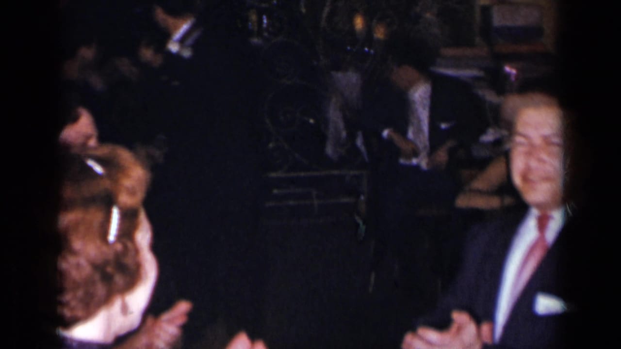Guests in formal attire clap and dance in a dimly lit crowded room.