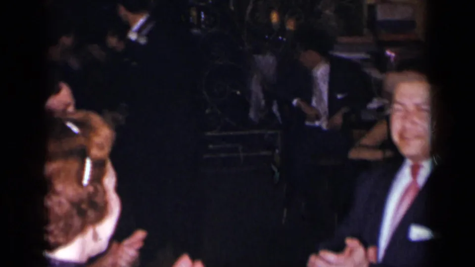1954 New York home-movie frame showing formally dressed adults clapping and dancing in a dark indoor party setting.