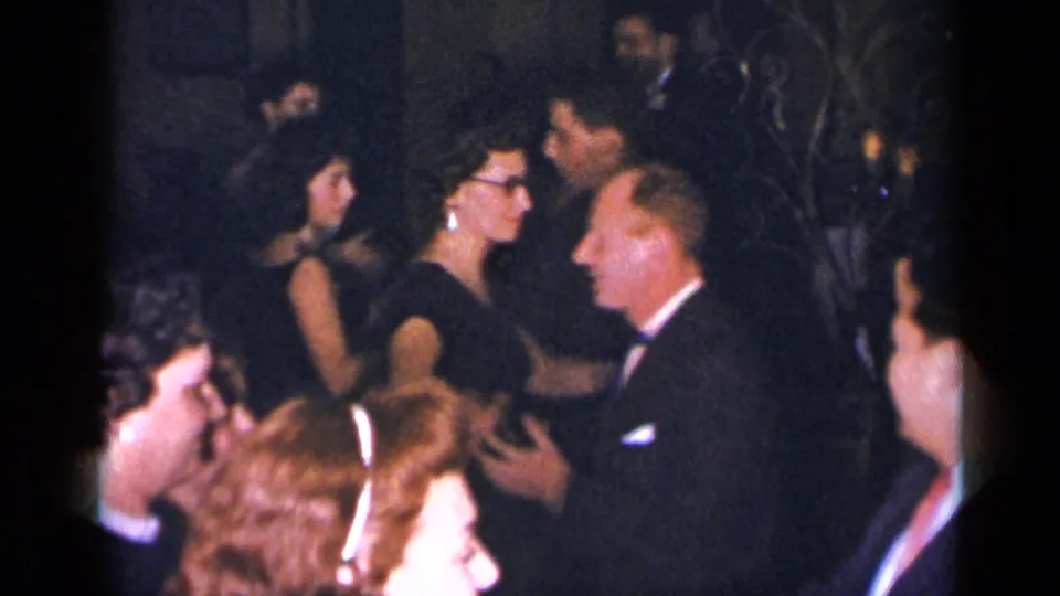1954 New York home-movie frame showing party guests dancing closely in formal dress inside a dim private room.