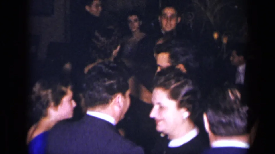 1954 New York party frame with adults in formal clothing dancing and conversing in a packed interior.