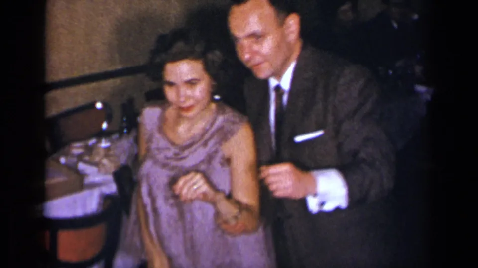 1954 New York home-movie frame of a woman in a light purple dress dancing with a suited man near set tables.