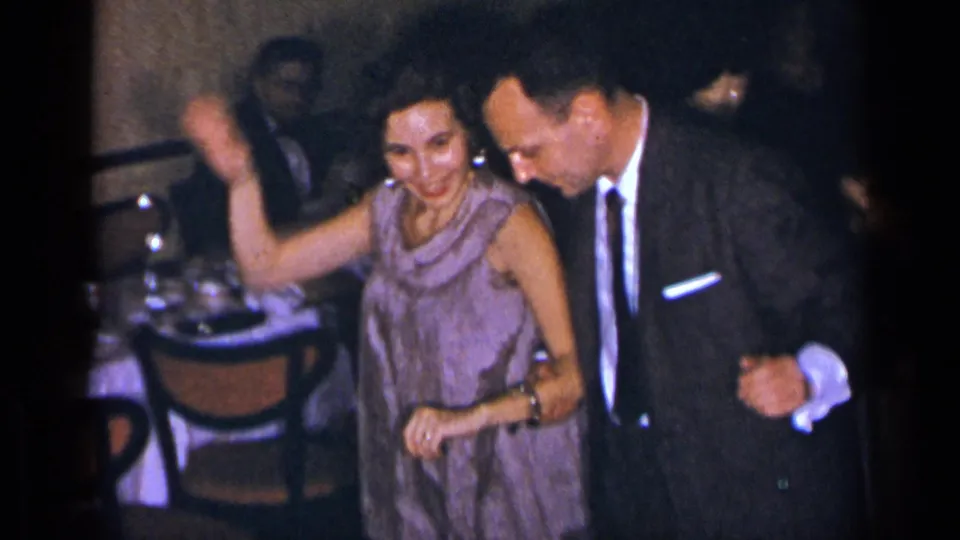 1954 New York formal party frame showing a smiling woman in a light purple dress dancing beside a man in a dark suit.