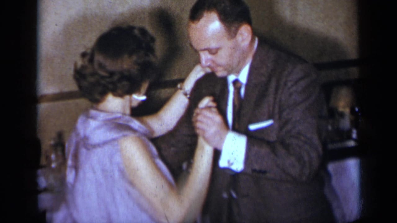 Man in a suit and woman in a lavender dress dance closely indoors.