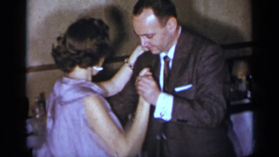 At a 1954 New York party, a suited man and woman in a lavender sleeveless dress hold hands while dancing closely indoors.