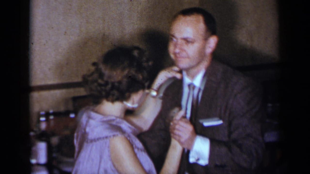 Woman faces a suited man as they hold hands while dancing indoors.
