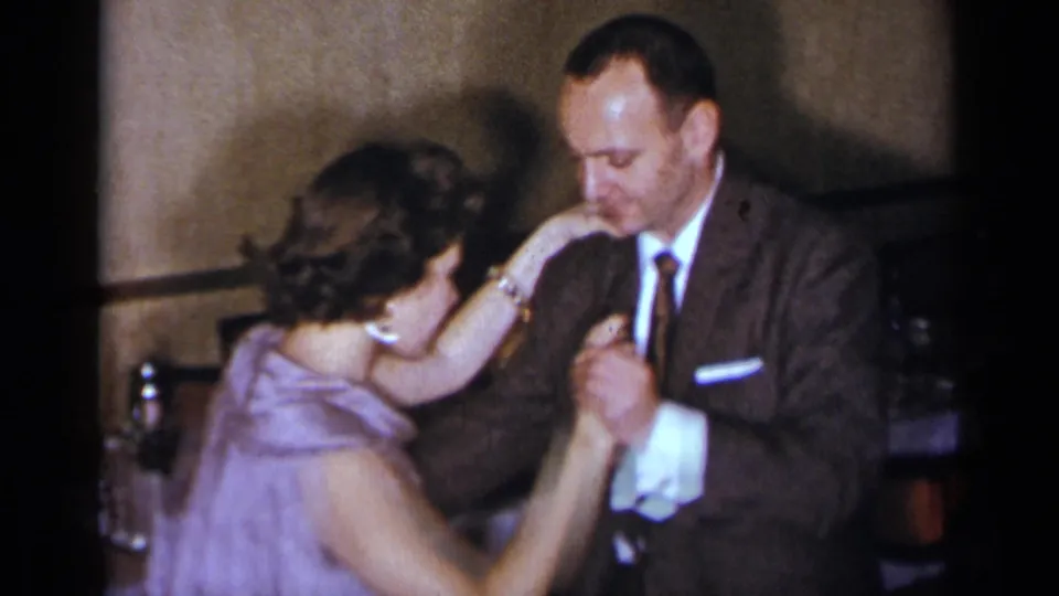 During the close dance, the suited man leans toward the woman's raised hand in a warmly lit 1954 New York party scene.