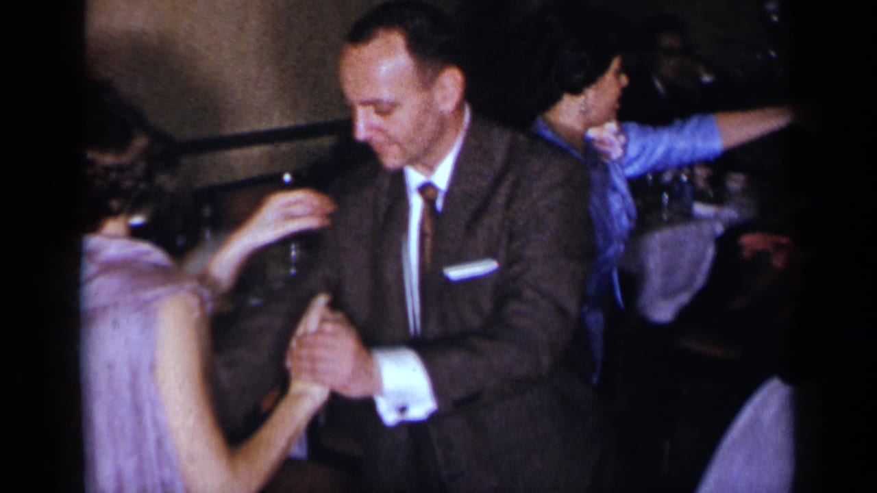 In 1954 New York home-movie footage, a suited man holds hands with a woman in a light dress while dancing at an indoor gathering.