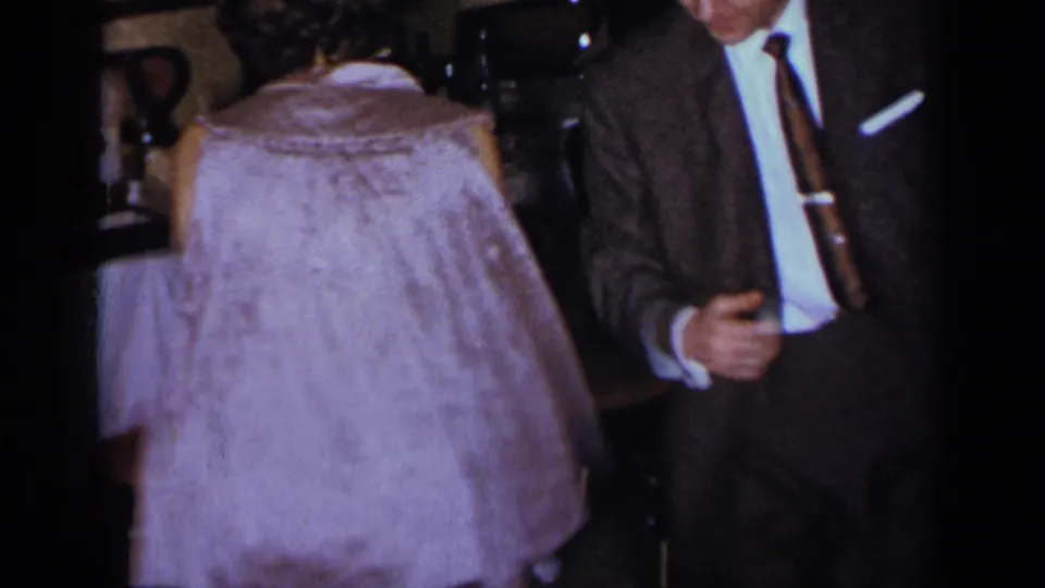 The woman’s light dress is seen from behind as she dances beside a suited man in soft, grainy home-movie footage.