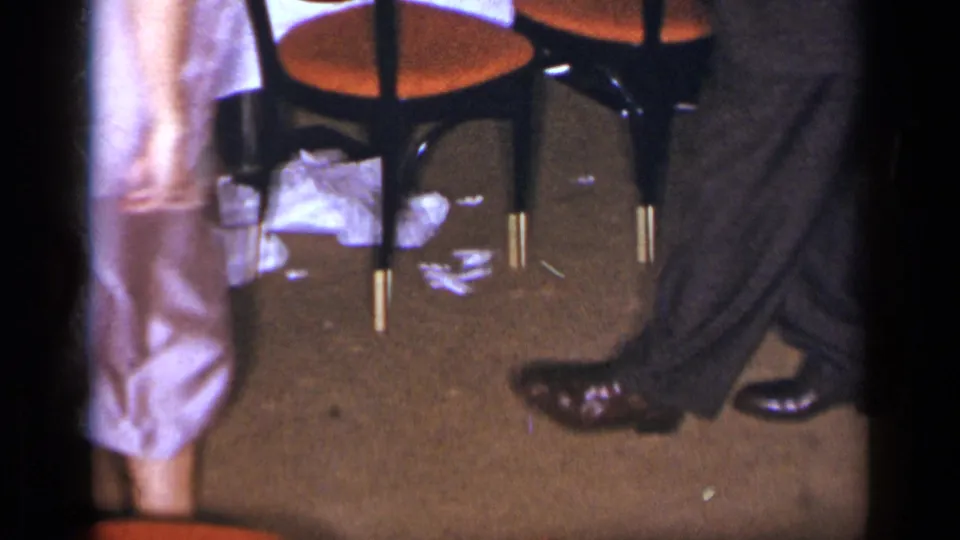 The dancers’ legs and shoes move across a carpeted private interior, with chairs and loose items visible nearby.