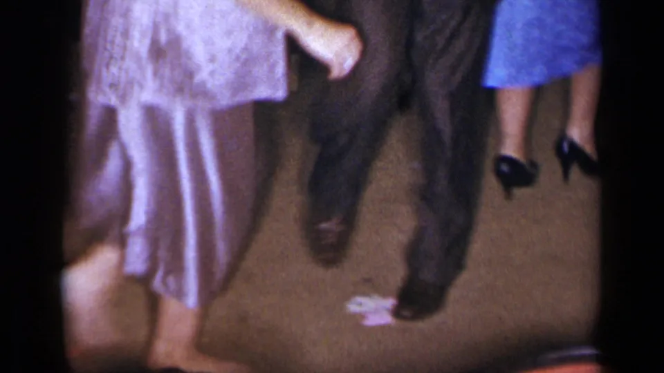 1954 New York home-movie frame showing adults dancing, including a light pleated dress, dark suit trousers, and dress shoes on an indoor floor.
