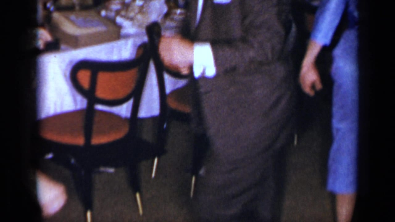 1954 New York indoor gathering frame with a suited man, a woman in a blue dress, a white tablecloth, and a black chair with an orange seat.
