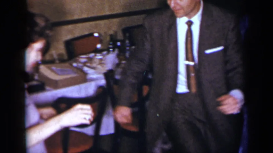 1954 New York home-movie frame showing a suited man dancing beside a table with chairs, tableware, and a partially visible seated person.