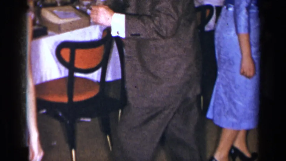 1954 New York social-gathering frame with a suited man, a woman in a blue dress, a white table, and an orange-seat chair in a hall interior.