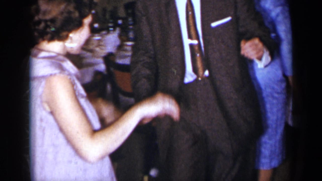 Woman in a light sleeveless dress dances near a suited man in a dim room.