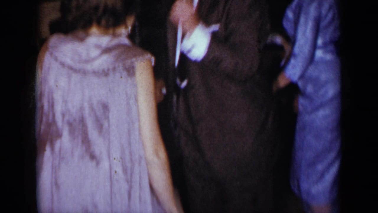 1954 New York home-movie frame with a woman in a light dress seen from behind among suited guests in a dim private room.