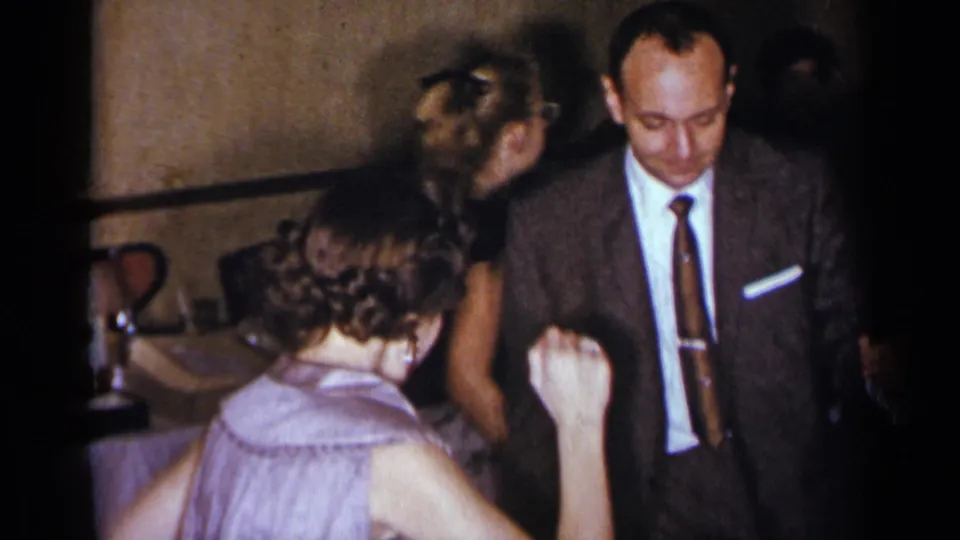 1954 New York home-movie frame showing a smiling suited man and women dancing near a table with glasses in a private interior.
