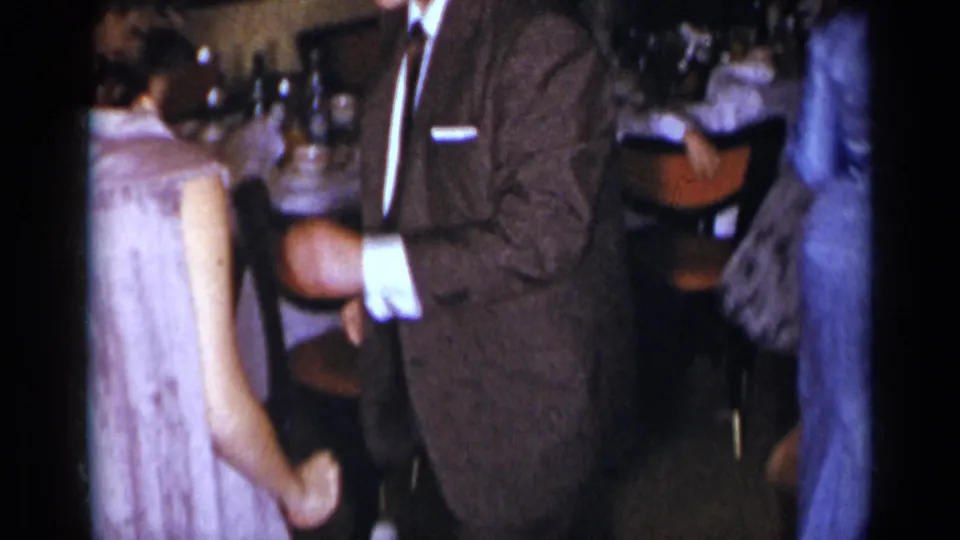1954 New York home-movie frame of a woman in a light sleeveless dress dancing close to a man in a suit in a dim indoor room.