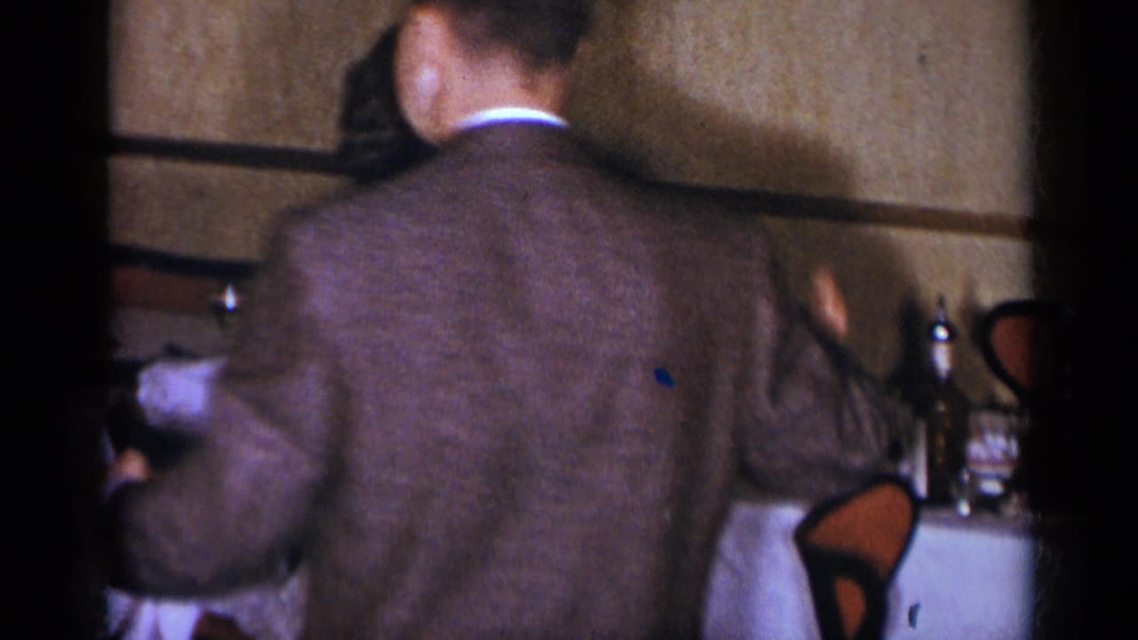 Man in a brown suit stands near a woman and a set dining table in a dim interior.