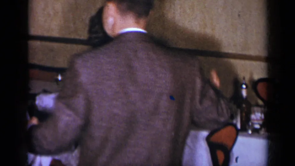 In 1954 New York home-movie footage, a man in a brown suit turns beside a woman and a table set with glassware and bottles.