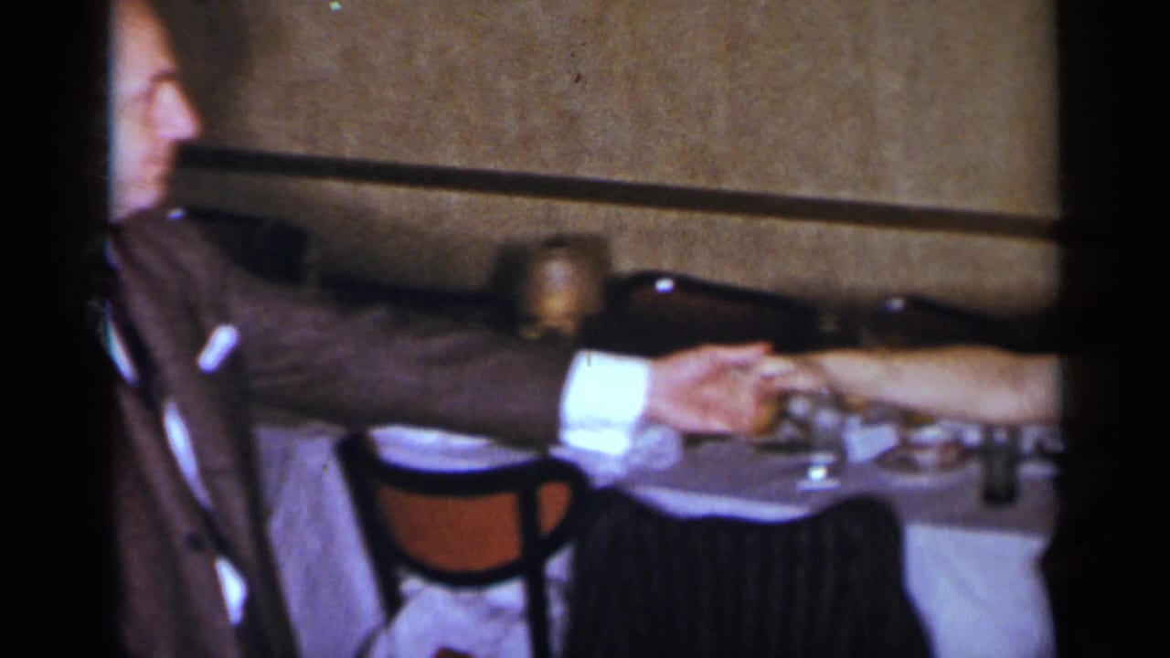 Man in a brown suit reaches toward another person across a formal dining table.