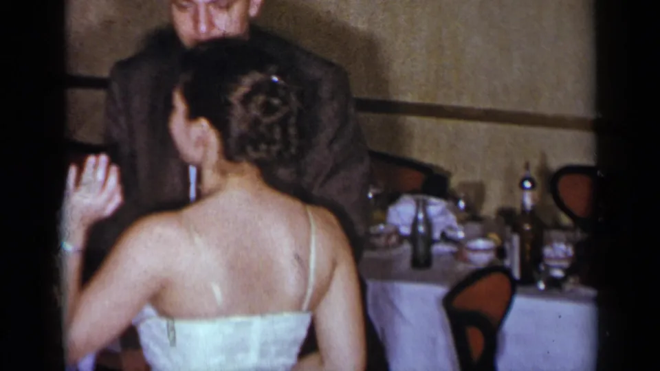 A woman in a white evening gown with an updo stands near a suited man and set table in 1954 New York home-movie footage.