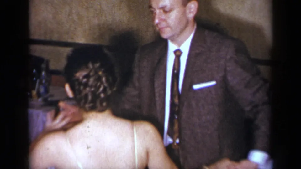 A suited man and a woman in a white gown face each other beside a formal table in a 1950s New York private interior.