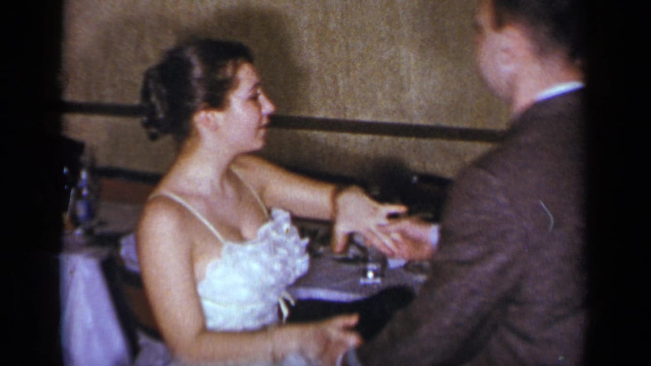 Woman in a light dress sits or leans near a table while holding a man's hand.