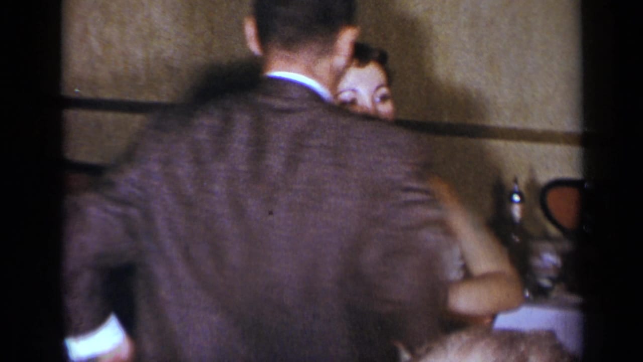 Man in a dark suit stands with his back to camera as a woman faces him near a table.